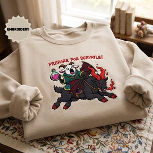 May include: A beige crewneck sweatshirt featuring an embroidered graphic of a white goat in a green robe holding a pink potion, riding a black winged creature with red horns and flames. The text above reads, "PREPARE FOR BEEYATLE!"