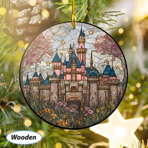 May include: Round wooden Christmas ornament featuring a castle design with pink, blue, and gray tones. The castle is surrounded by flowers and trees, with a stained-glass effect. The ornament is hanging from a gold string.