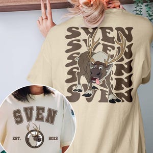 Pode incluir: Camiseta bege com um desenho de rena de desenho animado e o nome "SVEN" repetido. A frente da camiseta mostra "SVEN", um gr&aacute;fico de rena e o texto "EST. 2013".