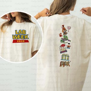 May include: Cream-colored t-shirt with the text "LAB WEEK 2026" in blue and yellow, with a red banner. The back of the shirt features colorful science-themed graphics, including a microscope and DNA.