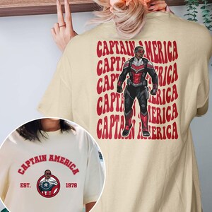 May include: Cream-colored t-shirt featuring a Captain America design. The front has a circular graphic with the text "Captain America" and "Est. 1978." The back displays a red and black Captain America illustration with the words "Captain America" repeated.