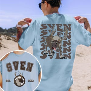 Pode incluir: Camiseta azul clara com uma rena de desenho animado chamada Sven e o nome "Sven" em letras marrons. A rena tem grandes chifres e a l&iacute;ngua de fora. A camisa tamb&eacute;m tem o texto "EST. 2013".