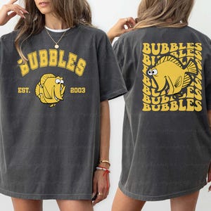 May include: Dark gray t-shirt with a yellow "BUBBLES" design and a cartoon fish. The front of the shirt has "EST. 2003" and a fish graphic. The back features a fish graphic and the word "BUBBLES" repeated.