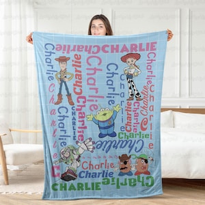 May include: A light blue fleece blanket featuring a grid pattern and the name Charlie repeated in various colors and fonts. The blanket displays cartoon characters including Woody, Jessie, an alien, Buzz Lightyear, and Mr. Potato Head.
