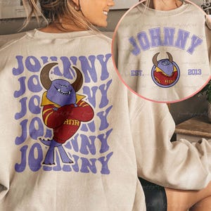 May include: Beige crew neck sweatshirt featuring a purple "JOHNNY" design and a cartoon bull mascot. The mascot is purple with horns, wearing a red and gold jacket. The back of the sweatshirt repeats the "JOHNNY" text.