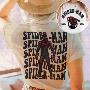 May include: A white t-shirt featuring a black and red Spider-Man graphic. The graphic is repeated several times in a retro style font. The text "SPIDER-MAN" is also printed on the shirt in a retro style font.