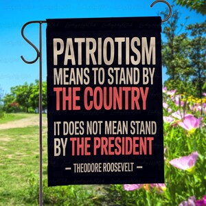 Puede incluir: Una bandera de jard&iacute;n negra con la cita "PATRIOTISMO SIGNIFICA APOYAR AL PA&Iacute;S, NO SIGNIFICA APOYAR AL PRESIDENTE - THEODORE ROOSEVELT -" en letras blancas y rojas. La bandera est&aacute; colgada de un poste de metal.