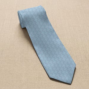 May include: A light blue necktie with a repeating wave pattern in a lighter shade of blue. The tie is made of a smooth fabric and is displayed on a neutral-colored surface.