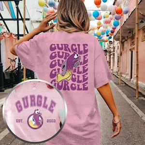 May include: Pink t-shirt with a purple fish graphic and the word "GURGLE" repeated in a retro font. The shirt also features a smaller graphic of the fish with the text "GURGLE EST. 2003" on a white circle.