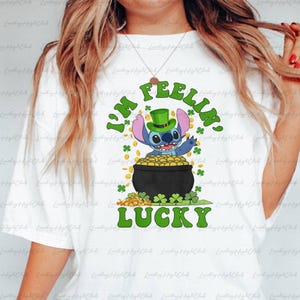 Puede incluir: Camiseta blanca con un personaje de dibujos animados con un sombrero verde de duende y el texto "I'M FEELIN' LUCKY". El personaje está sentado en una olla de monedas de oro, rodeado de tréboles verdes.