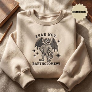 May include: A beige crewneck sweatshirt featuring an embroidered graphic of a grey gargoyle with wings. The text above the gargoyle reads FEAR NOT, and the text below reads BARTHOLOMEW!, accompanied by small embroidered star accents.