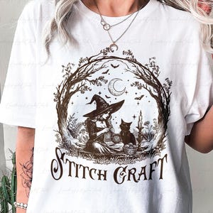 May include: A white t-shirt featuring a brown graphic print of a witch wearing a pointed hat, sitting and knitting with a black cat nearby. The scene is framed by tree branches under a crescent moon and stars, with the text STITCH CRAFT below.