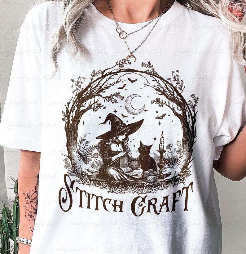 May include: A white t-shirt featuring a brown graphic print of a witch wearing a pointed hat, sitting and knitting with a black cat nearby. The scene is framed by tree branches under a crescent moon and stars, with the text STITCH CRAFT below.