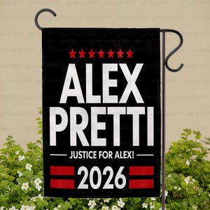 May include: Black garden flag with white text that reads "ALEX PRETTI" above "JUSTICE FOR ALEX!" and "2026". The flag has red stars and stripes. The flag is hanging on a black metal stand.