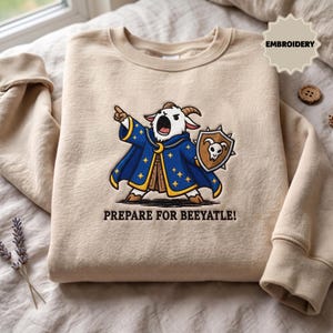 May include: A beige crewneck sweatshirt featuring an embroidered graphic of a white goat dressed as a wizard in a blue star-patterned robe, holding a shield with a skull emblem. The text below the goat reads, "PREPARE FOR BEEYATLE!" in black.