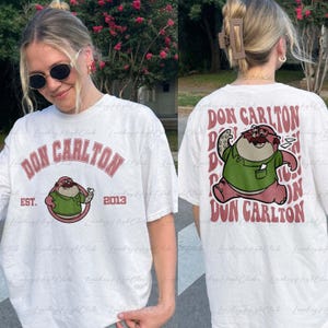 May include: White t-shirt featuring a cartoon character wearing a green shirt and holding a piece of paper. The text "DON CARLTON" is printed on the shirt in pink letters. The text "EST. 2013" is printed below the character.