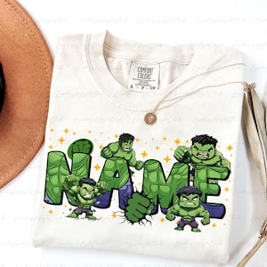 May include: Cream-colored t-shirt featuring the word "NAME" in a green, Hulk-themed design. The design includes illustrations of the Hulk and yellow star accents. A gold necklace and a brown hat are also visible.