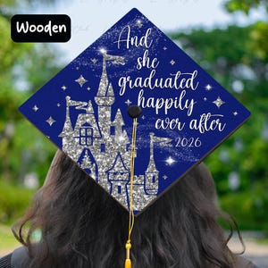 May include: A navy blue graduation cap with a silver glitter castle design and the text "And she graduated happily ever after 2026". The cap has a gold tassel and is made of wood.