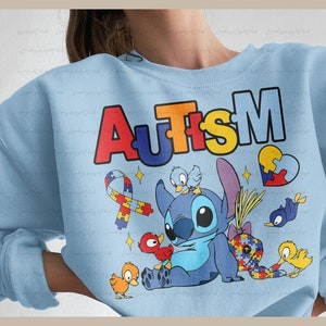 Op de afbeelding: Lichtblauwe trui met het woord "AUTISME" in regenboogkleuren. Een blauw Stitch-personage uit Lilo & Stitch bevindt zich in het midden van het ontwerp, omringd door kleine vogels en puzzelstukjes.