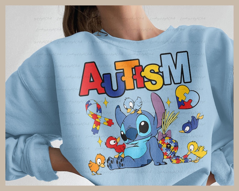 Op de afbeelding: Lichtblauwe trui met het woord "AUTISME" in regenboogkleuren. Een blauw Stitch-personage uit Lilo & Stitch bevindt zich in het midden van het ontwerp, omringd door kleine vogels en puzzelstukjes.