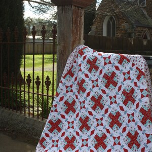 May include: A red and white quilt with a geometric pattern. The quilt is folded and draped over a wooden post in front of a stone building.