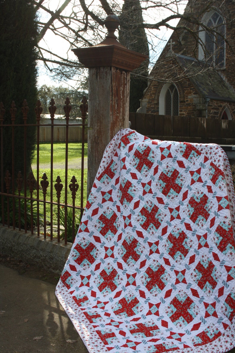 May include: A red and white quilt with a geometric pattern. The quilt is folded and draped over a wooden post in front of a stone building.