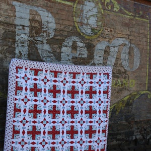 May include: A red, white, and blue quilt with a geometric pattern. The quilt is hanging on a brick wall with a faded sign that says "Reg.