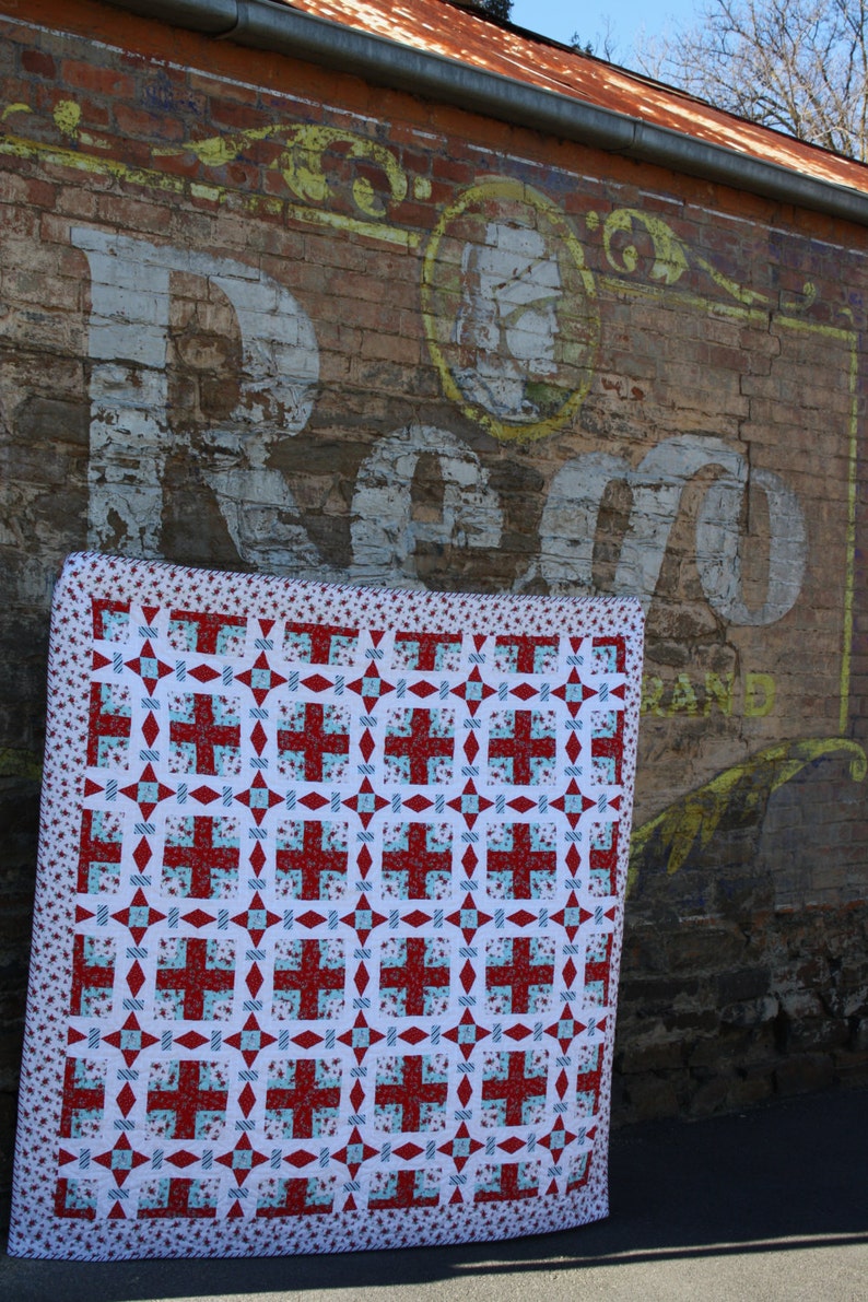 May include: A red, white, and blue quilt with a geometric pattern. The quilt is hanging on a brick wall with a faded sign that says "Reg.