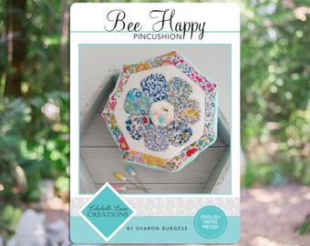 May include: A colorful floral pincushion in the shape of a flower with a bee in the center. The pincushion is made using the English paper piecing technique. The pincushion is on a light wood surface. The text "Bee Happy Pincushion" is at the top of the image. The text "Libelbelle Lane Creations" is at the bottom left of the image. The text "By Sharon Burgess" is below the text "Libelbelle Lane Creations". The text "English Paper Pieced" is in a circle at the bottom right of the image.