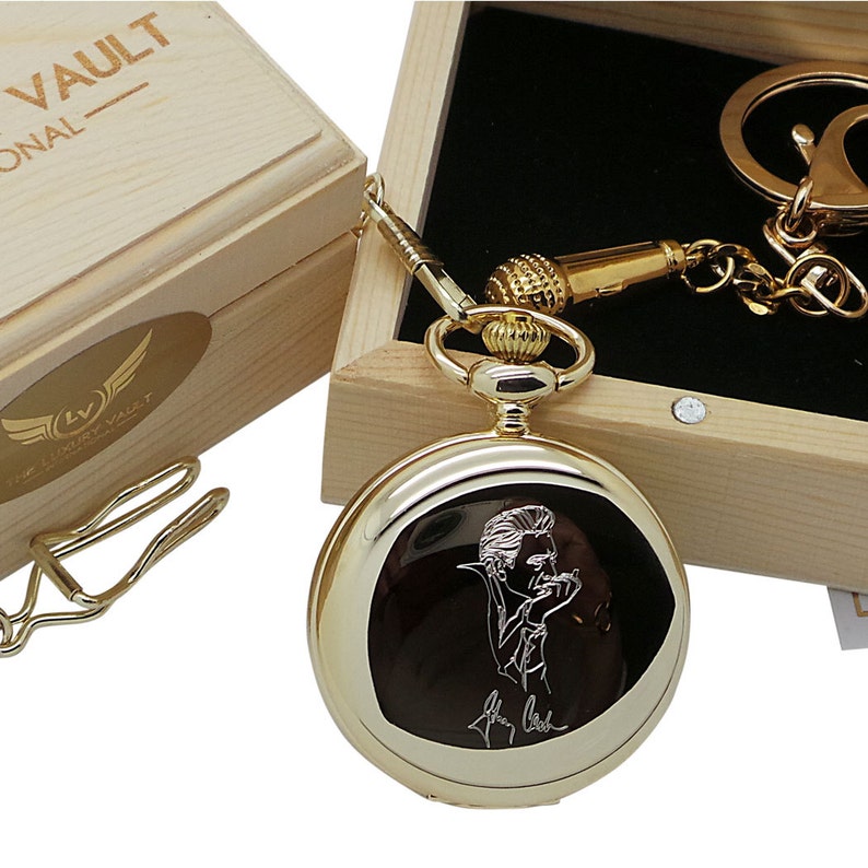 May include: Gold pocket watch with a black face and a white engraved image of a man holding a microphone. The watch is in a wooden box with a gold chain and a microphone charm.