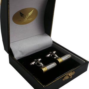 Op de afbeelding: Een paar zilveren en gouden kogel manchetknopen in een zwarte geschenkdoos met een grijs interieur. De doos heeft een gouden logo met de tekst "The Luxury Vault International".