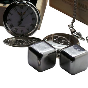 May include: A silver pocket watch with a white face and black Roman numeral hour markers. The watch is open and the back is visible. Two silver metal cubes with the initials "KS" engraved on them are in the foreground.