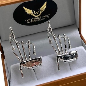 Puede incluir: Un par de gemelos plateados con forma de gaita, presentados en una caja de madera con interior blanco. La caja presenta una etiqueta ovalada negra con alas doradas y el texto "THE LUXURY VAULT INTERNATIONAL".