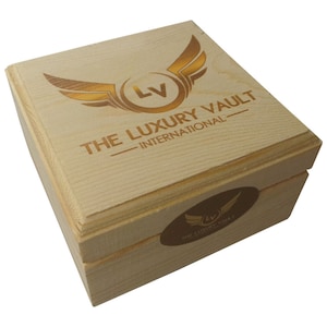 May include: A wooden box with a gold logo that says "The Luxury Vault - International" and a smaller logo on the side. The box is made of light wood and has a natural finish.