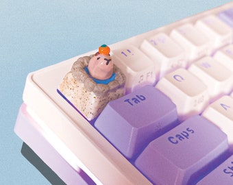 May include: A close-up of a keyboard with a custom keycap featuring a small, pink animal figurine wearing a blue hat and an orange fruit. The keyboard has purple keys with white lettering and a white base.