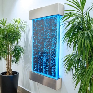 May include: A contemporary wall-mounted water feature with a stainless steel frame. The clear acrylic panel is lit with blue LED lights, producing a bubble effect. Potted green plants are positioned on either side, adding a natural element to the interior design.