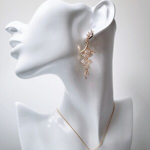 May include: A gold-toned metal earring featuring a tiered, fan-shaped design with clear baguette-cut stones, paired with a matching gold-toned chain necklace that has a single, small, dangling, fan-shaped pendant set with clear baguette-cut stones.