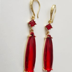 Pode incluir: Um par de brincos pendurados dourados com pedras preciosas vermelhas em forma de gota. Cada brinco apresenta uma pequena gema vermelha quadrada no topo, conectada a uma gema vermelha maior e alongada. Os brincos têm um gancho decorativo.