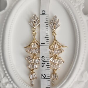 May include: A pair of gold-toned dangle earrings featuring clear baguette-cut crystals arranged in a tiered fan design. A white measuring tape is placed vertically between the earrings, showing a total length of approximately 2.25 inches.