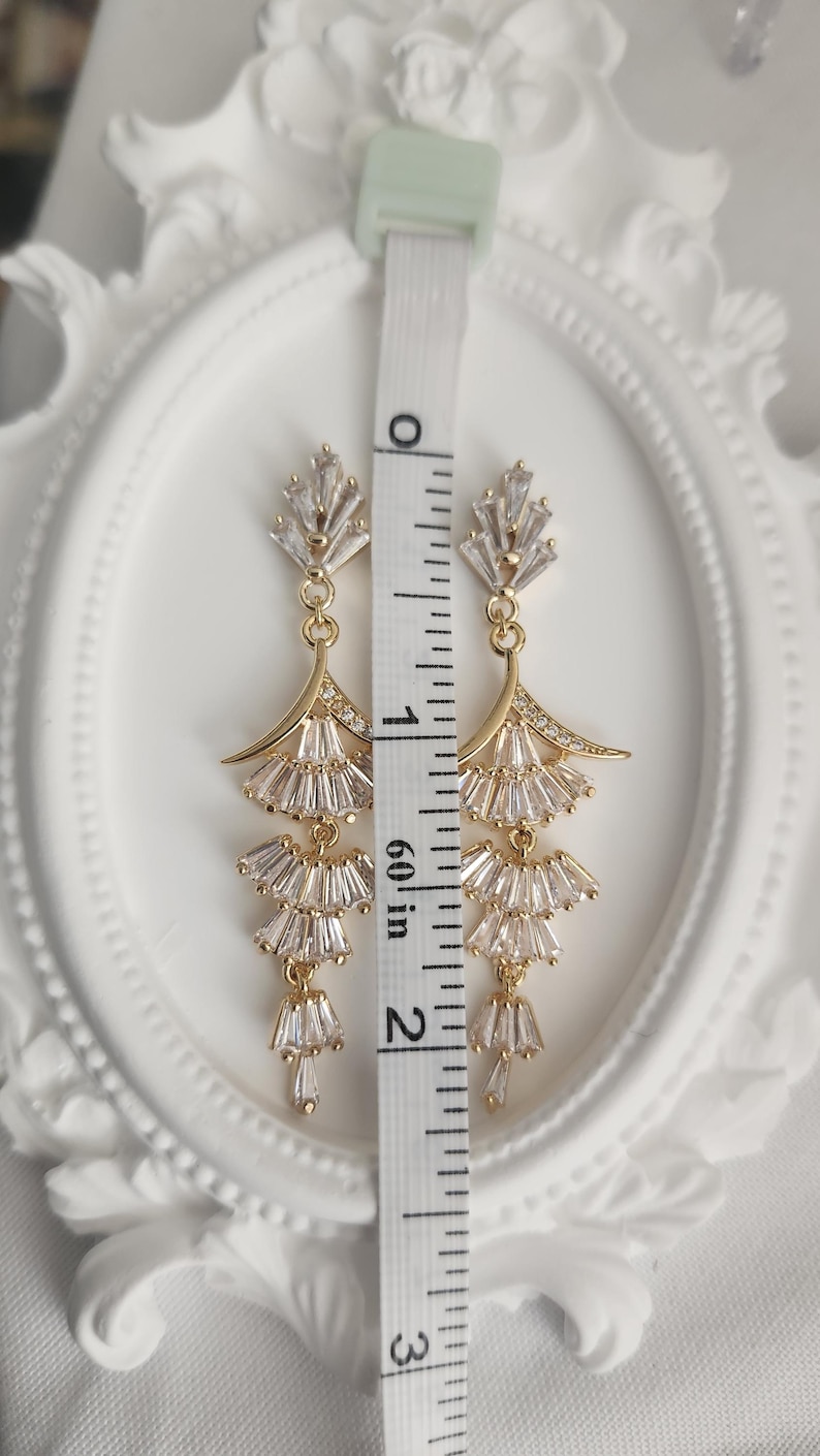 May include: A pair of gold-toned dangle earrings featuring clear baguette-cut crystals arranged in a tiered fan design. A white measuring tape is placed vertically between the earrings, showing a total length of approximately 2.25 inches.