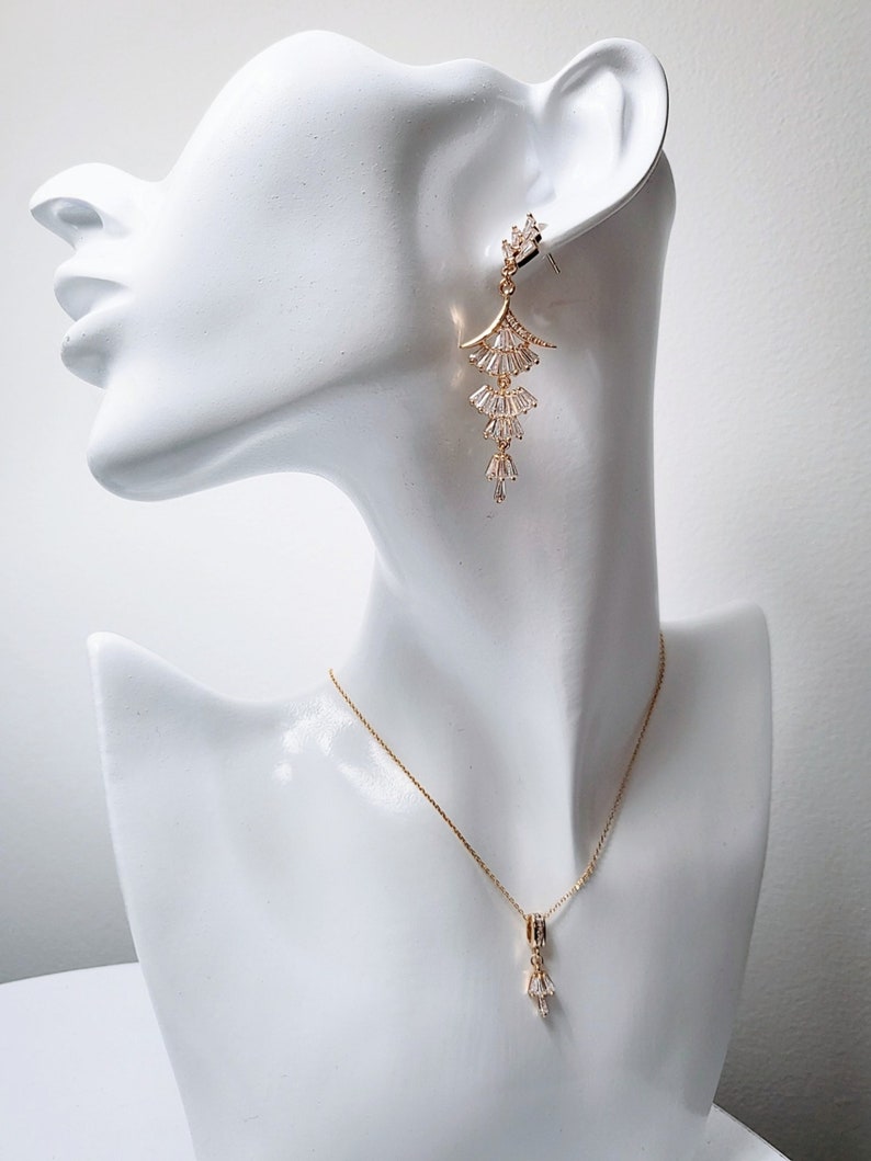 May include: A gold-toned metal earring featuring a tiered, fan-shaped design with clear baguette-cut stones, paired with a matching gold-toned chain necklace that has a single, small, dangling, fan-shaped pendant set with clear baguette-cut stones.