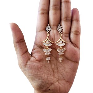May include: A pair of gold-toned dangle earrings featuring a tiered, fan-like design. Each earring is adorned with multiple clear, baguette-cut crystals arranged in a cascading pattern, suspended from a small, triangular crystal stud top.