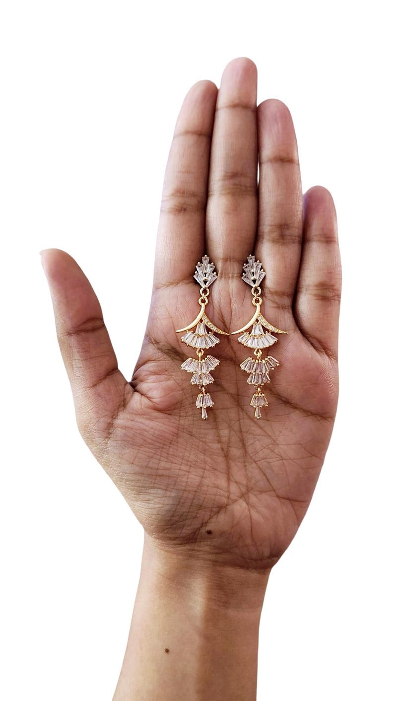 May include: A pair of gold-toned dangle earrings featuring a tiered, fan-like design. Each earring is adorned with multiple clear, baguette-cut crystals arranged in a cascading pattern, suspended from a small, triangular crystal stud top.