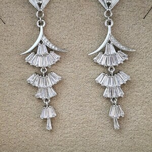 May include: A pair of silver-toned drop earrings featuring a tiered design. Each earring consists of a top fan-shaped element followed by three descending, smaller fan-shaped sections, all encrusted with clear baguette-cut crystals.