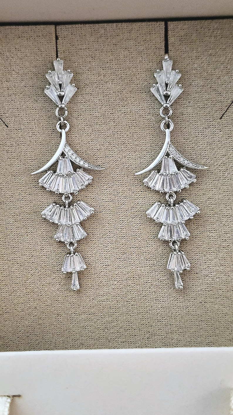 May include: A pair of silver-toned drop earrings featuring a tiered design. Each earring consists of a top fan-shaped element followed by three descending, smaller fan-shaped sections, all encrusted with clear baguette-cut crystals.