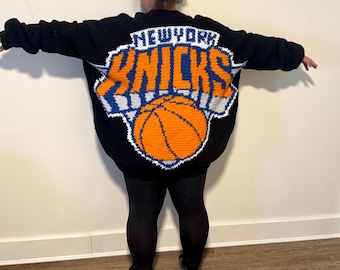 May include: Black knitted sweater featuring the New York Knicks logo in orange, blue, and white. The logo includes the team name and a basketball. The sweater is oversized and has long sleeves.