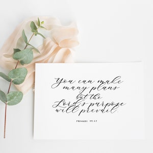 Könnte beinhalten: Eine weiße Karte mit schwarzer Kalligrafie, auf der steht: "Du kannst viele Pläne machen, aber der Plan des Herrn wird sich durchsetzen." Der Bibelvers "Sprüche 19:21" ist unter dem Text gedruckt. Die Karte liegt auf einer weißen Oberfläche mit einem Zweig Eukalyptus und einem hellrosa Band.