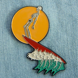 May include: Enamel pin featuring a skeleton surfing on a red surfboard in front of an orange sun. The wave is white and green. The pin is set against a blue denim background.