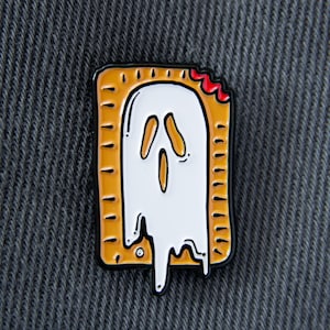 Puede incluir: Pin esmaltado con un diseño de fantasma dentro de una Pop-Tart. El fantasma es blanco con un efecto de goteo, y la Pop-Tart tiene una corteza marrón dorada con un mordisco, revelando un relleno rojo. El pin tiene un contorno negro.