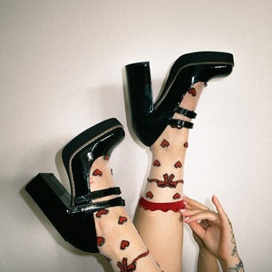 May include: A pair of black platform shoes with a buckle strap and a red heart patterned sheer sock. The shoes have a thick platform and a high heel.